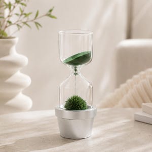 May include: A magnetic hourglass featuring a clear glass vessel with a silver-colored base. Inside the glass, dark green magnetic sand forms a spiky, spherical shape at the bottom, while a small amount of green sand remains at the top.