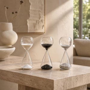 May include: Three clear glass hourglasses arranged in a row. The left hourglass contains white sand, the centre hourglass contains black sand, and the right hourglass contains grey sand. Each hourglass features a rounded top and a conical base.