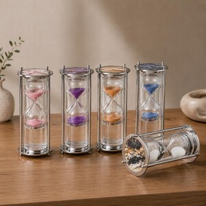 May include: Five cylindrical hourglasses with metallic frames and marble-patterned tops. Four stand upright containing pink, purple, orange, and blue sand, while one lies horizontally featuring white sand and a black and white marble top.