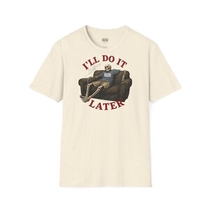 Può includere: Una t-shirt a maniche corte color crema con la grafica di uno scheletro che si rilassa su un divano marrone scuro. Il testo "I'LL DO IT LATER" è stampato in lettere rosse arcuate sopra e sotto l'illustrazione. Lo scheletro indossa una maglietta blu.