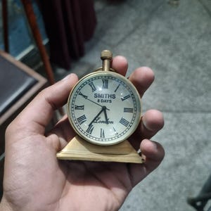 Puede incluir: Un pequeño reloj de color latón con esfera redonda y números romanos. La esfera del reloj dice "SMITHS 8 DAYS London". El reloj se sostiene en una mano y tiene una pequeña base rectangular.