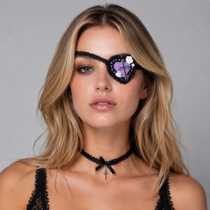 May include: A heart-shaped eye patch featuring a purple center, a black lace border, a silver cross charm, and small white floral and heart accents. A matching black lace choker with a thin ribbon bow and a small silver cross pendant is worn.