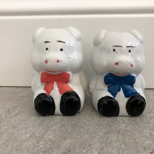 May include: Two white ceramic piggy banks with painted details. One piggy bank has a red bow and the other has a blue bow.