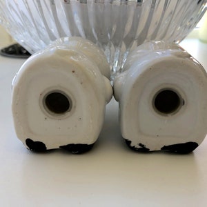 May include: A pair of white ceramic salt and pepper shakers shaped like a couple of people holding hands. The shakers have black painted details on the bottom.