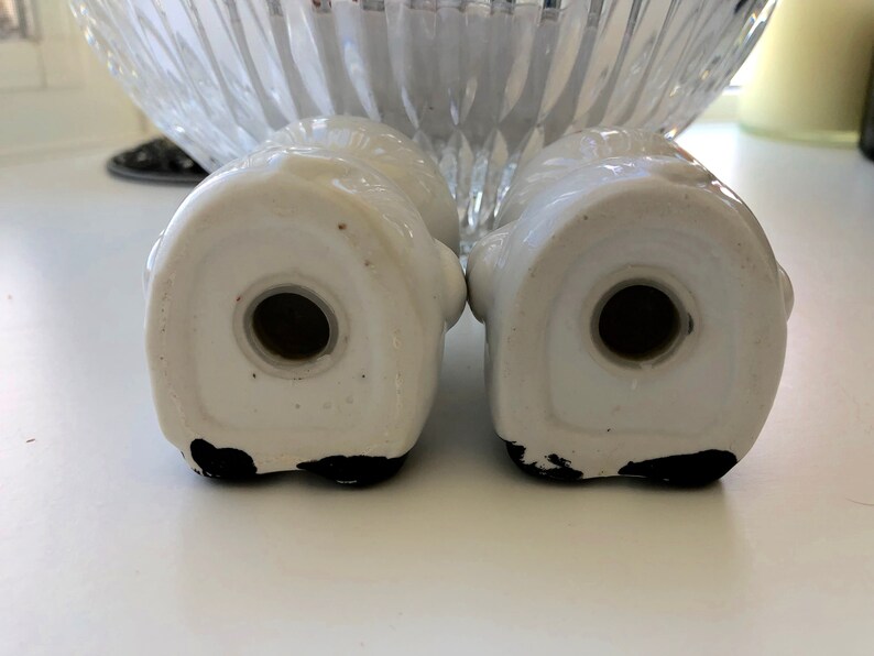 May include: A pair of white ceramic salt and pepper shakers shaped like a couple of people holding hands. The shakers have black painted details on the bottom.