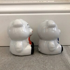May include: Two white ceramic piggy banks, one with a red bow and the other with a blue bow. The piggy banks are facing each other.