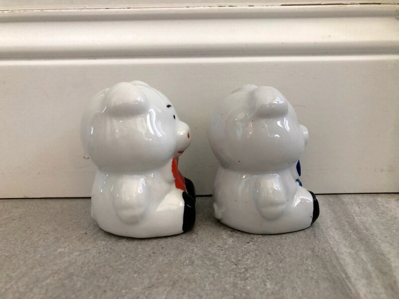 May include: Two white ceramic piggy banks, one with a red bow and the other with a blue bow. The piggy banks are facing each other.