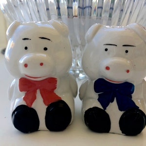 May include: Two white ceramic pig salt and pepper shakers. The pig on the left has a red bow tie and the pig on the right has a blue bow tie.