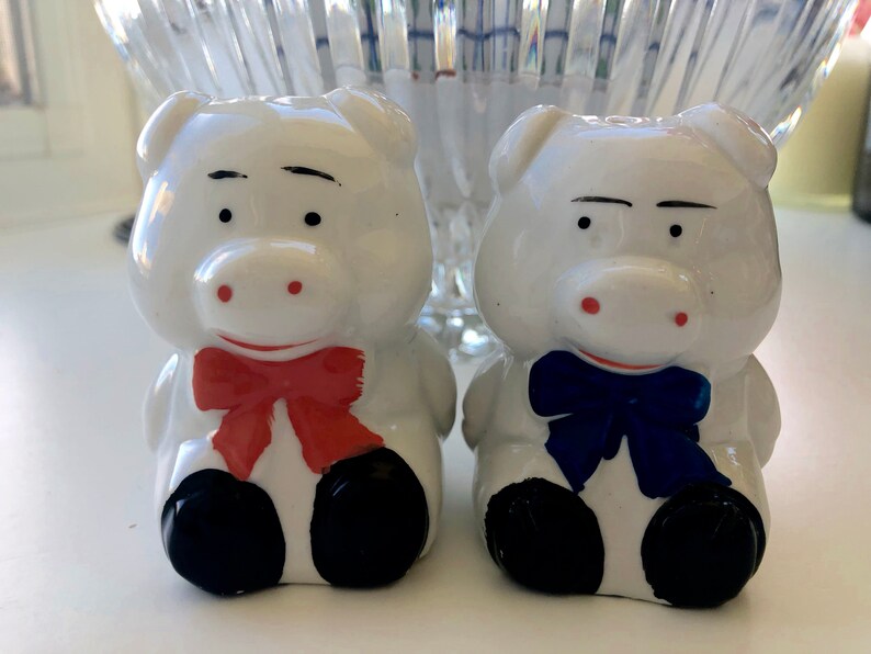 May include: Two white ceramic pig salt and pepper shakers. The pig on the left has a red bow tie and the pig on the right has a blue bow tie.
