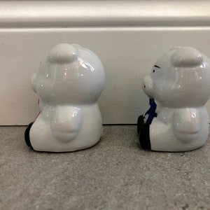 May include: Two white ceramic piggy banks, one with a red bow and the other with a blue bow. The piggy banks are sitting on a gray surface.