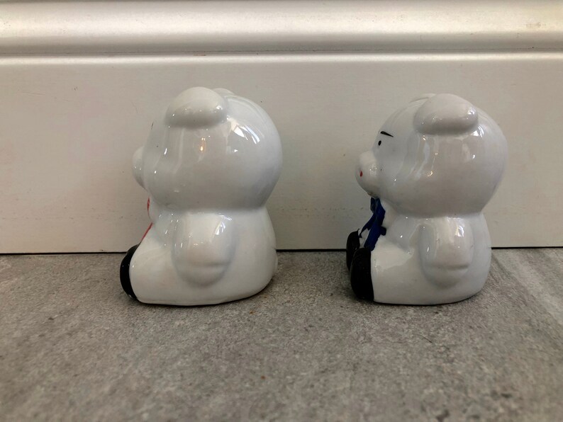 May include: Two white ceramic piggy banks, one with a red bow and the other with a blue bow. The piggy banks are sitting on a gray surface.