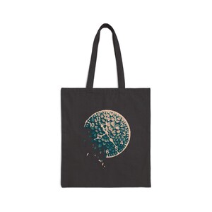 May include: A black tote bag featuring a graphic print of a clock face composed of dark teal and light beige petal-like shapes. The clock design appears to be disintegrating on the left side, with small fragments floating away from the main circular form.