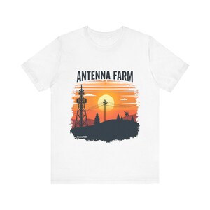 Peut inclure: T-shirt blanc avec un motif de coucher de soleil représentant une silhouette de tour d'antenne, des arbres et le texte "ANTENNA FARM". Le graphisme est en orange, noir et blanc.