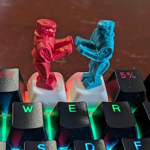 May include: Two small robot figurines, one red and one blue, standing on white mechanical keyboard keycaps. The keyboard features black keys with illuminated green and red characters, including the numbers 2, 5, and 6, and letters W, E, R, T.