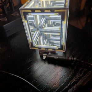 May include: A square infinity mirror cube featuring internal LED light strips along each edge. The device has a clear acrylic frame, a reflective interior creating a tunnel effect, and a base with a power port labeled DC5V and a small white switch.