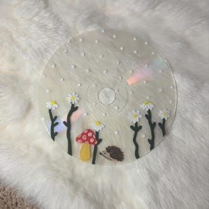 May include: A clear CD with a hand-painted design featuring a red mushroom with white spots, daisies, and a hedgehog. The daisies have white petals and yellow centers. The background has white dots.