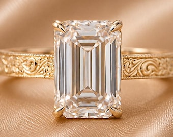 May include: A gold ring featuring a large, rectangular emerald-cut clear gemstone held by four prongs. The band is crafted from yellow gold and displays an intricate, embossed floral scrollwork pattern along the entire outer surface of the shank.