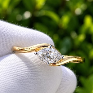 May include: A gold ring featuring a marquise-cut diamond set in a white metal prong setting. The band has a smooth, polished finish and a gentle wave-like design that tapers toward the center stone, creating a sleek and refined appearance.