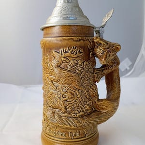 以下が含まれることがあります： 鹿と狐のレリーフデザインが施された茶色の陶器製ビアジョッキ。持ち手は狐の形をしており、蓋は銀色の金属製です。ジョッキの底面にはDie grüne Heidという浮き彫りの文字が刻まれています。