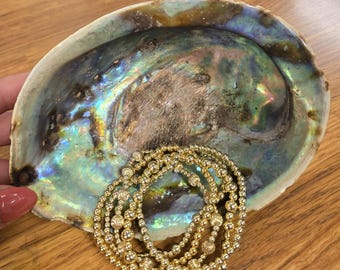 May include: Abalone shell with iridescent colors, holding a collection of gold beaded bracelets. The bracelets are made of small, round beads and are arranged in a layered design. The shell sits on a wooden surface.