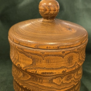 Pode incluir: Um pote de madeira artesanal com tampa. O pote é de cor castanha quente com padrões de grão de madeira visíveis. A tampa tem um pequeno botão arredondado. O corpo do pote apresenta um design decorativo em camadas. A estética geral é rústica e natural.