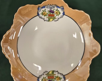 May include: A vintage, scalloped-edge dish with a cream-colored interior and a peach-colored rim. The rim features a black border and is decorated with floral designs in yellow, purple, and blue. The dish is likely made of porcelain.