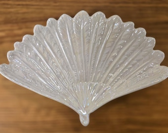 May include: Iridescent white glass fan-shaped dish. The dish has a scalloped edge and a textured, feather-like pattern. The dish is on a wooden surface.