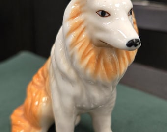May include: A ceramic figurine of a Collie dog in a sitting position. The dog has a white body with orange-brown fur around the neck and ears. The dog's eyes are dark, and the nose is black. The figurine is on a green surface.