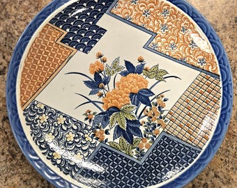 May include: A round ceramic plate with a blue rim and a floral design. The plate features a central floral arrangement in orange and blue, surrounded by geometric patterns in blue and orange. The plate's rim has a wave-like pattern.