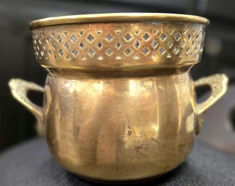May include: A vintage brass pot with a decorative pierced rim and two ornate handles. The pot has a rounded body and a warm, golden hue, suggesting age and use. The rim features a pattern of diamond-shaped cutouts.