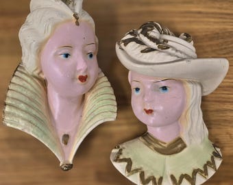 May include: Two vintage ceramic wall plaques depicting female faces. One wears a feathered headdress, the other a wide-brimmed hat. Both have pale pink skin, blue eyes, and red lips. The plaques feature gold and light green accents.