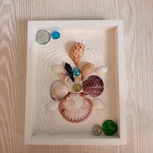 Könnte beinhalten: Ein weiß gerahmtes Kunstwerk mit Muscheln und Glasperlen. Das Kunstwerk zeigt verschiedene Muscheln in Braun-, Weiß- und Burgundertönen auf einem strukturierten weißen Hintergrund. Blaue und grüne Glasperlen setzen farbliche Akzente.