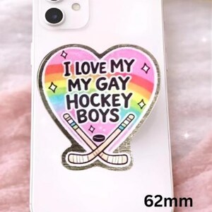 Op de afbeelding: Een witte telefoon met een sticker met de tekst "I LOVE MY GAY HOCKEY BOYS" in een hartvorm met een regenboog en hockeysticks. De sticker is 6,2 cm groot.