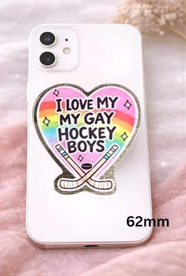 Op de afbeelding: Een witte telefoon met een sticker met de tekst "I LOVE MY GAY HOCKEY BOYS" in een hartvorm met een regenboog en hockeysticks. De sticker is 6,2 cm groot.