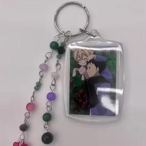 May include: A clear acrylic keychain featuring an anime-style image of two characters. The keychain has a silver ring and a beaded charm with pink, green, purple, and black beads, and a red flower-shaped charm.
