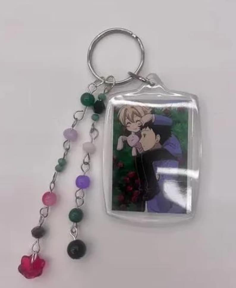 May include: A clear acrylic keychain featuring an anime-style image of two characters. The keychain has a silver ring and a beaded charm with pink, green, purple, and black beads, and a red flower-shaped charm.