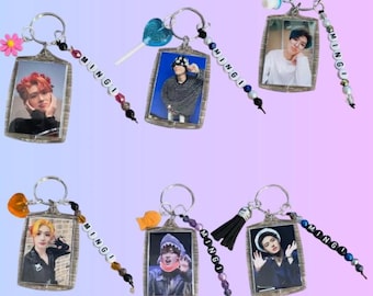 May include: A collection of keychains featuring rectangular photo frames with images of people. Each keychain has a silver ring, a beaded charm with the name "MINGI", and decorative accents like flowers, lollipops, and tassels. The background is a gradient of purple and blue.