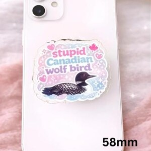 Op de afbeelding: Een witte smartphone met een decoratieve telefoonhouder. De houder heeft een cartoonillustratie van een ijsvogel met de tekst "stupid Canadian wolf bird" en een esdoornblad. De houder meet 5,8 cm.