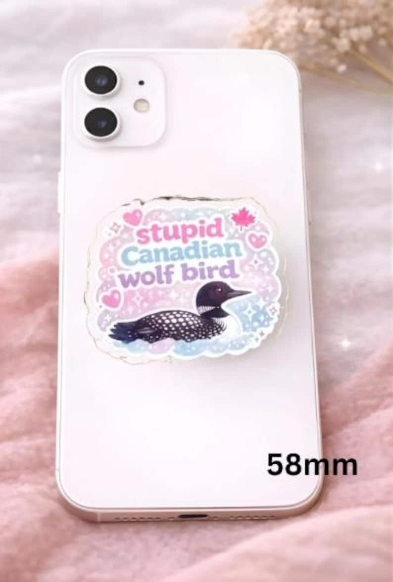 Op de afbeelding: Een witte smartphone met een decoratieve telefoonhouder. De houder heeft een cartoonillustratie van een ijsvogel met de tekst "stupid Canadian wolf bird" en een esdoornblad. De houder meet 5,8 cm.