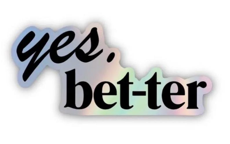 May include: A holographic sticker with the words "yes. bet-ter" in black script. The sticker has a white border and a shimmering, iridescent finish, reflecting a spectrum of colours.