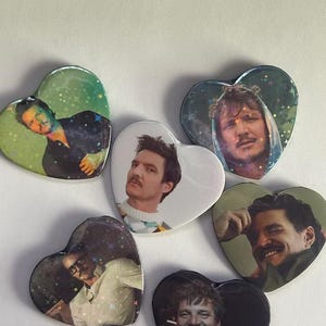 May include: Five heart-shaped pinback buttons featuring various photographic portraits of a man with a mustache. Four of the buttons have a holographic star overlay, while the central button has a plain white background.
