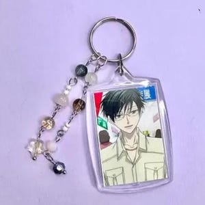 May include: Clear acrylic keychain featuring an anime character with black hair and glasses. The keychain has a silver keyring and a beaded charm with various colored beads and a star-shaped charm.