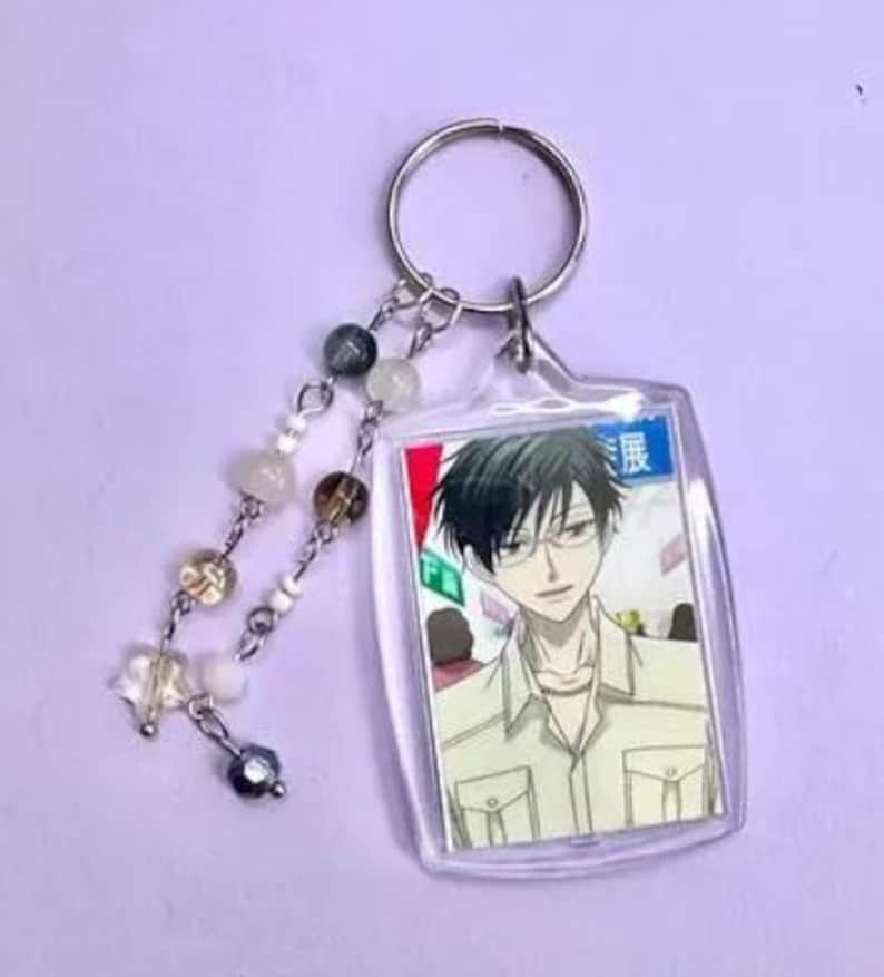 May include: Clear acrylic keychain featuring an anime character with black hair and glasses. The keychain has a silver keyring and a beaded charm with various colored beads and a star-shaped charm.