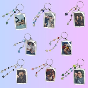 May include: Eight rectangular clear acrylic keychains, each featuring a unique photograph of people. Every keychain is attached to a metal ring adorned with a dangling chain of colorful beads, including small stars, spheres, and translucent accents.