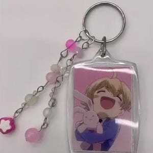 May include: A clear acrylic keychain featuring a cartoon image of a smiling character holding a stuffed animal. The keychain has a silver ring and a beaded charm with pink and white beads and a pink flower-shaped bead.