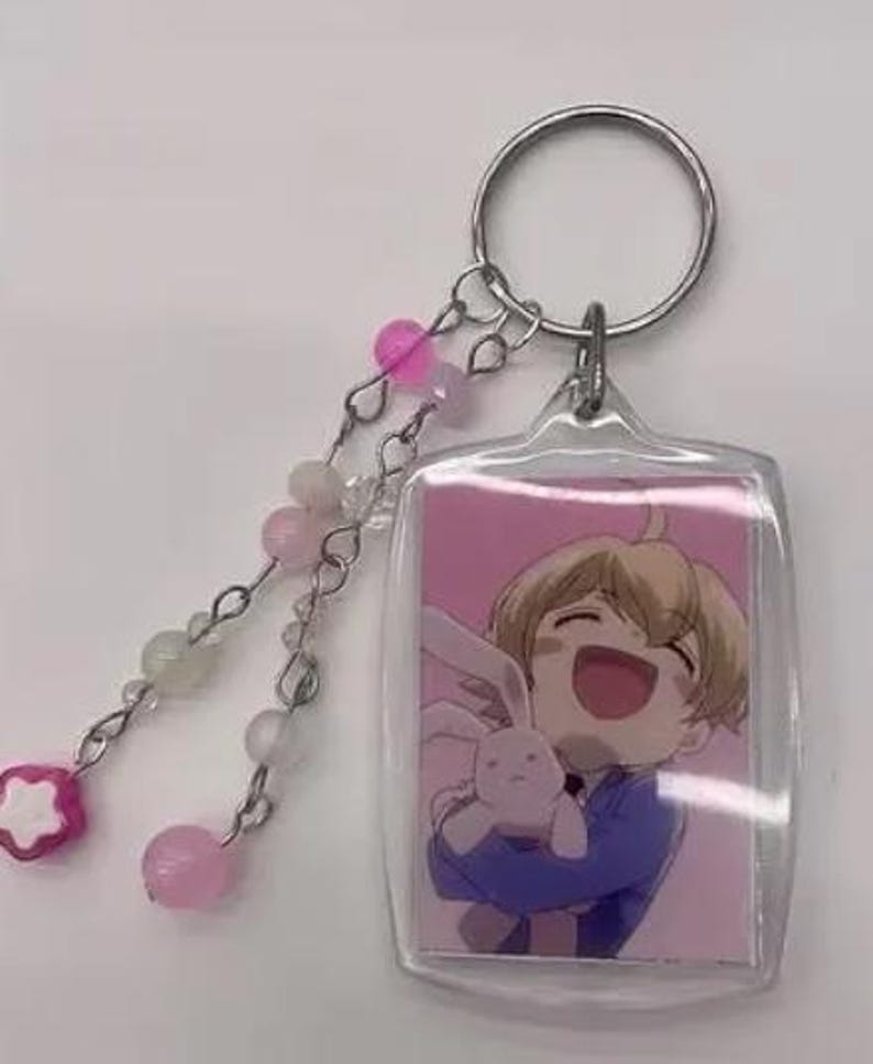 May include: A clear acrylic keychain featuring a cartoon image of a smiling character holding a stuffed animal. The keychain has a silver ring and a beaded charm with pink and white beads and a pink flower-shaped bead.