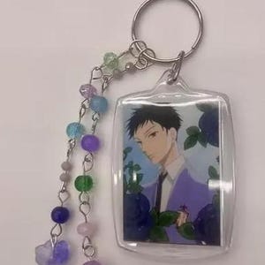 May include: Clear acrylic keychain featuring an anime-style character in a purple suit, surrounded by blue flowers. The keychain has a silver keyring and a beaded charm with various colored beads, including purple, blue, and green.