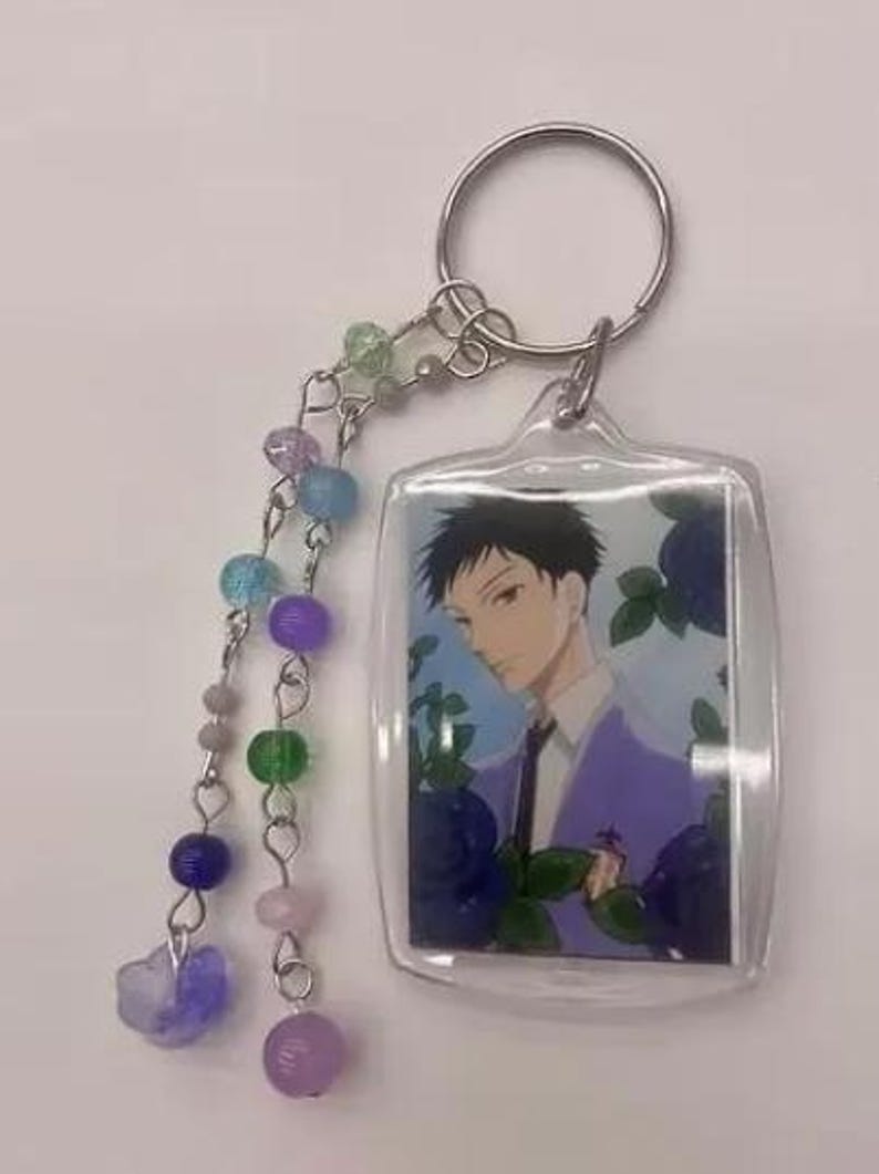 May include: Clear acrylic keychain featuring an anime-style character in a purple suit, surrounded by blue flowers. The keychain has a silver keyring and a beaded charm with various colored beads, including purple, blue, and green.