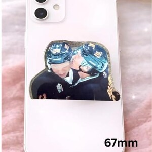 Op de afbeelding: Witte smartphone met een decoratieve sticker met twee hockeyspelers in zwarte uniformen en helmen, de een de ander kussend. De sticker heeft een gouden rand. De telefoon is 6,7 cm lang.