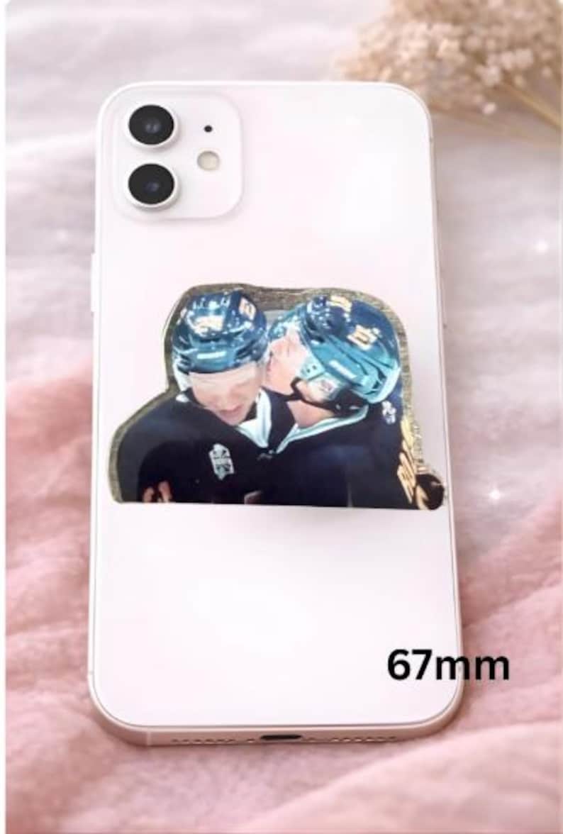 Op de afbeelding: Witte smartphone met een decoratieve sticker met twee hockeyspelers in zwarte uniformen en helmen, de een de ander kussend. De sticker heeft een gouden rand. De telefoon is 6,7 cm lang.